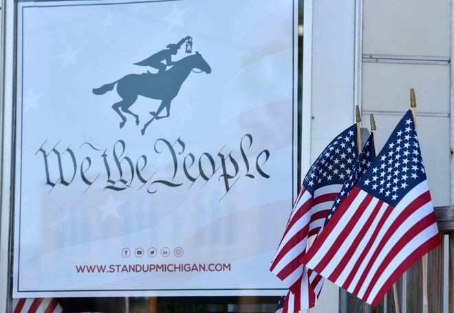 A poster of a horse and rider, representing Paul Revere with the words, 'We the People' is displayed near three U.S. flags in the barbershop window. This is sponsored by StandUpMichigan.com, which is a movement to get Michigan businesses back open.
