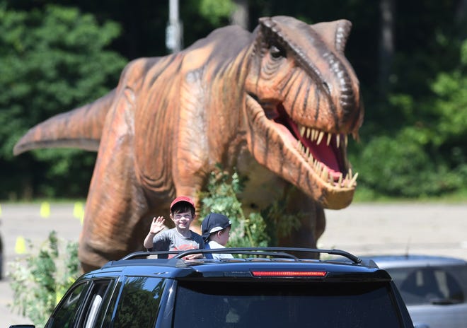 Archie Steigleman waves at dinosaurs with his brother Louie as they approach the mighty Tyrannosaurus Rex which towers over vehicles.